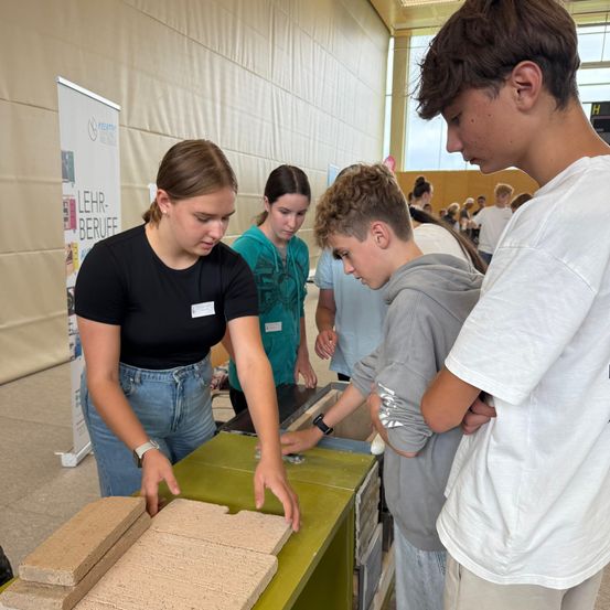 Eine Frau und mehrere junge Menschen untersuchen einen Stapel Ziegel. Sie sind in einem Klassenzimmer. Im Hintergrund sind andere Menschen und ein Banner zu sehen.