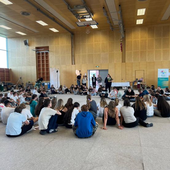 In einem großen, gut beleuchteten Raum sitzt eine Gruppe von Menschen im Kreis auf dem Boden. Mehrere Personen stehen in der Mitte. Einige halten Mikrofone, während andere beobachten. Banner und Ausrüstung sind an den Seitenlinien vorhanden.