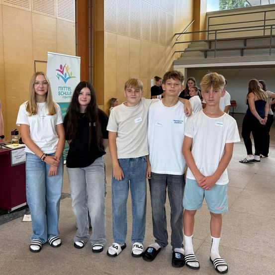 Fünf junge Menschen, drei Jungen und zwei Mädchen, posieren für ein Foto in einem Schulflur. Sie tragen Freizeitkleidung. Hinter ihnen befindet sich ein Banner mit Text und ein Tisch mit Gegenständen. In der Ferne stehen und sitzen Menschen auf den Stufen.