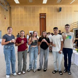 Fünf junge Menschen stehen in einem Raum und halten Getränke in den Händen. Im Hintergrund befindet sich ein Tisch mit Blumen.