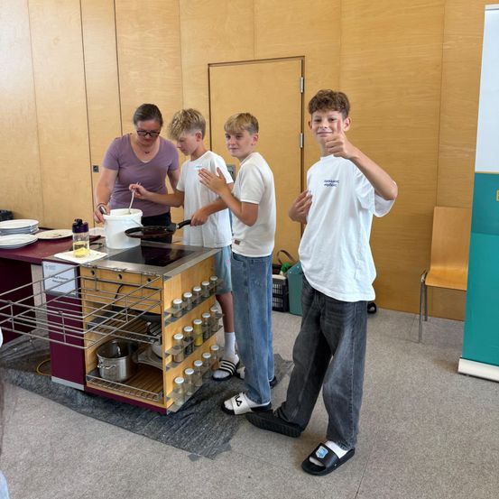 Eine Frau und drei Jungen stehen um einen Kochbereich. Die Frau rührt in einem Topf, während die Jungen zusehen. Ein Junge gibt einen Daumen hoch.