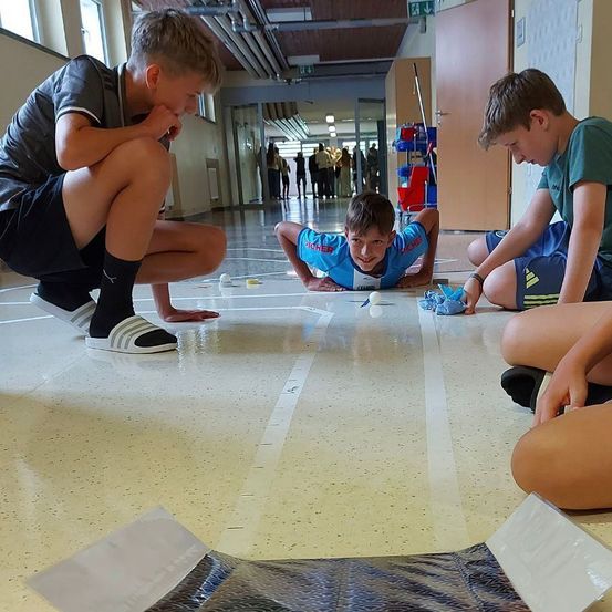 Drei Jungen sind in einem Spiel auf einem polierten Boden engagiert. Einer liegt auf dem Bauch, ein anderer hockt, und ein dritter kniet, alle konzentriert auf das Spiel.