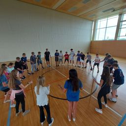 Eine Gruppe von Kindern und Erwachsenen übt in einer Turnhalle mit Holzboden einen Sport mit Stöcken. Sie sind in zwei Kreise unterteilt, einen innerhalb des anderen.