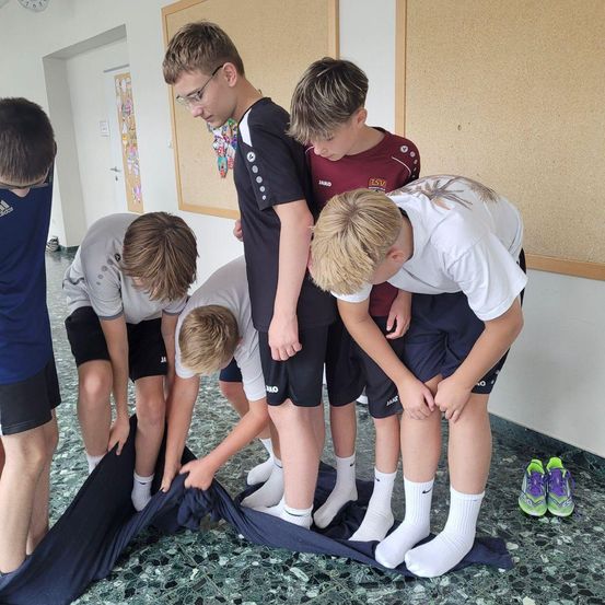 Eine Gruppe von Jungen steht um ein blaues Tuch herum und wechselt wahrscheinlich ihre Socken. Sie befinden sich in einem Raum mit gefliesten Böden und einer Uhr an der Wand.