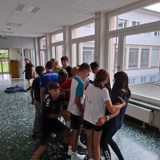 Eine Gruppe junger Leute steht in einem Raum mit großen Fenstern zusammen, lächelt und interagiert. Kleidung ist auf dem Boden verstreut.