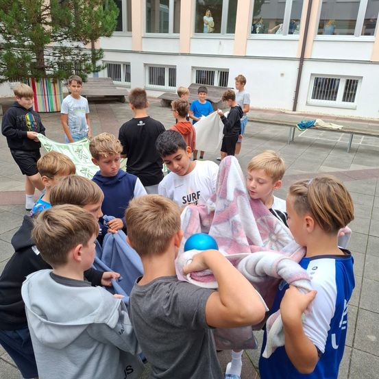 Eine Gruppe von Jungen in Freizeitkleidung, versammelt um ein Gebäude mit mehreren Fenstern, hält Handtücher und einen Ball und bereitet sich möglicherweise auf eine Aktivität oder ein Spiel vor.