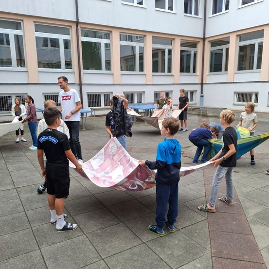 Kinder sind draußen in der Nähe eines Gebäudes, einige halten große Stoffbahnen. Ein Mann steht vorne und spricht. Ein anderer Junge hält ein farbenfrohes Stück Stoff.