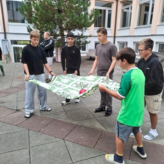 Sechs Jungen spielen mit einem grünen Netz. Ein Junge hält das Netz, während die anderen Jungen versuchen, den Ball zu fangen. Die Jungen tragen Freizeitkleidung. Dahinter befindet sich ein Gebäude mit Glasfenstern.