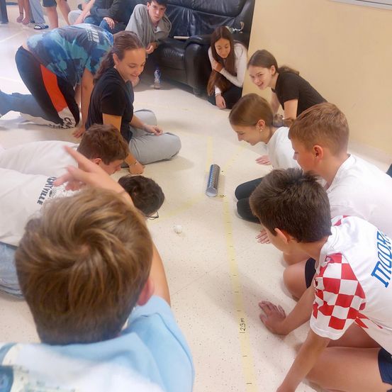 Eine Gruppe junger Menschen nimmt an einer Teambuilding-Aktivität auf dem Boden teil. Sie tragen verschiedene Shirts und schauen auf eine gelbe Linie und einen Ball.