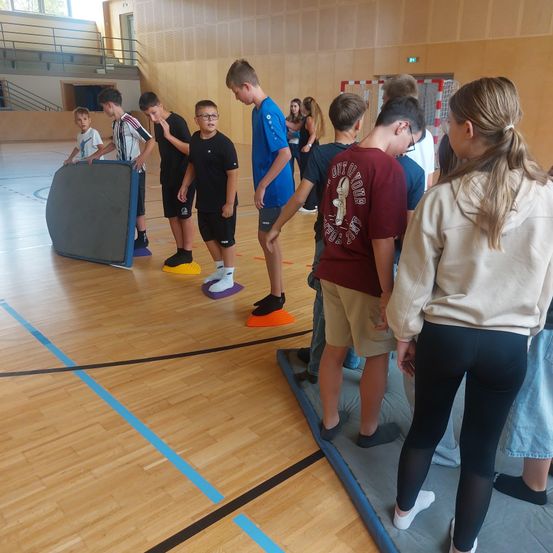 In einer Turnhalle sind eine Gruppe junger Menschen bei einer Aktivität, die auf verschiedenen farbigen Matten stehen. Sie tragen Turnschuhe, Brillen und Freizeitkleidung.