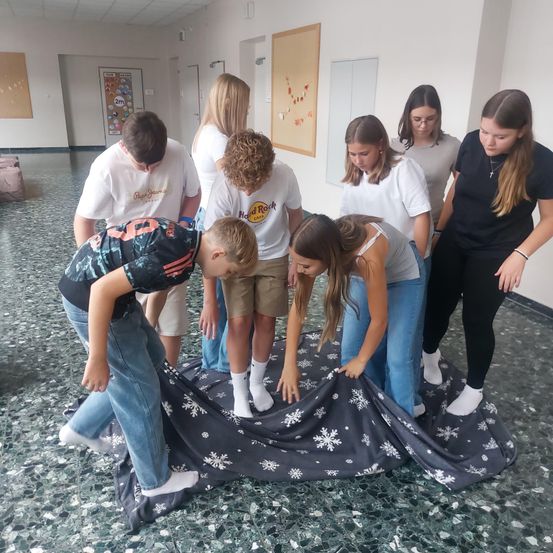 Eine Gruppe von Menschen befindet sich in einem Raum, sie beugen sich über eine Decke mit Schneeflocken. Sie tragen Freizeitkleidung. Hinter ihnen befinden sich Poster an der Wand und eine Tür.