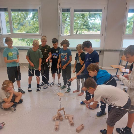 In einem Klassenzimmer beschäftigen sich eine Gruppe von Jungen mit einer Aktivität, die Schnüre und Holzblöcke beinhaltet. Sie stehen und sitzen um eine zentrale Struktur herum, wobei einige Schnüre halten. Der Raum hat große Fenster, die natürliches Licht hereinlassen.