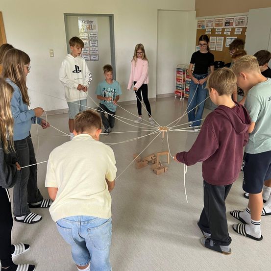 Schüler sind um einen zentralen Punkt herum versammelt, durch Seile verbunden und bilden eine spinnennetzartige Struktur mit Blöcken und einer Lehrerin im Hintergrund.