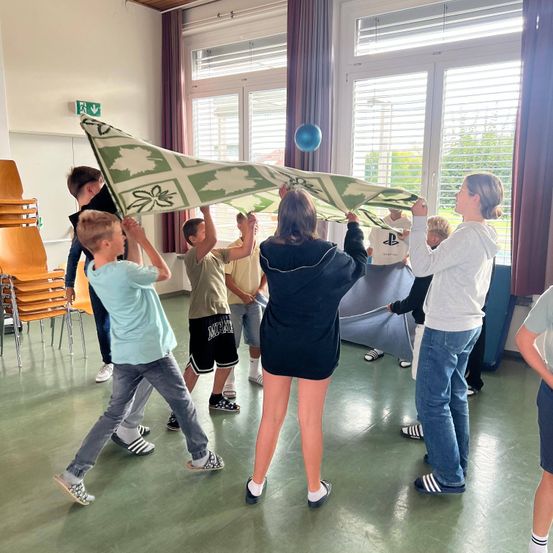 Eine Gruppe von Kindern und Erwachsenen versammelt sich in einem Raum und hält ein großes grünes Tuch mit Blattmustern. Sie bereiten sich auf eine Aktivität vor.