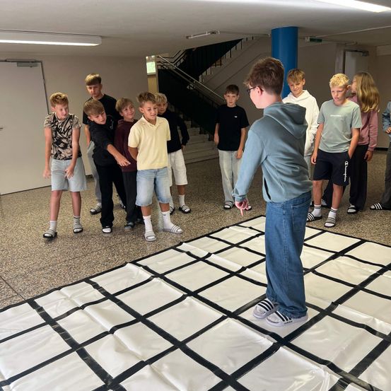 Eine Gruppe von Jungen und Mädchen in einem Raum, ein Junge steht auf einem Gittermatte, ein anderer Junge zeigt auf ihn, während andere zusehen.