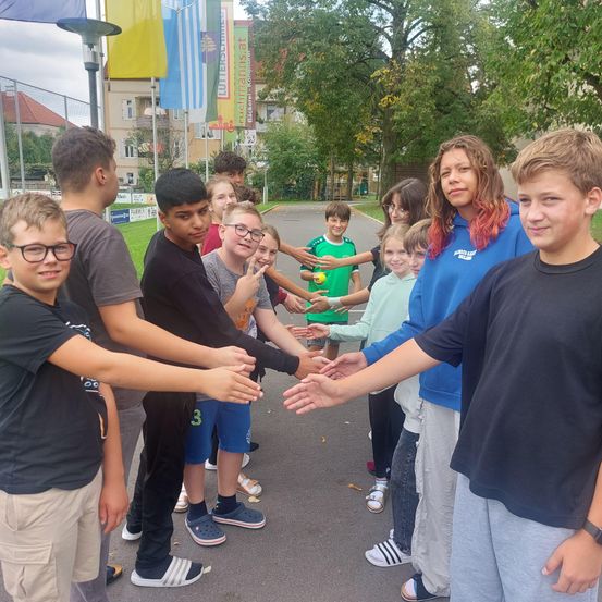 Eine Gruppe von Kindern, die sich an den Händen halten und in einer Reihe auf einer Straße stehen, mit Gebäuden und Bäumen im Hintergrund.
