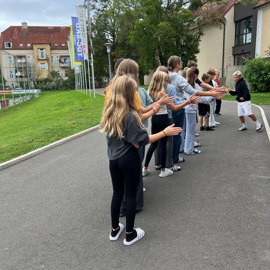 Eine Gruppe von Kindern und Jugendlichen steht lächelnd in einer Reihe auf einer Straße. Sie sind draußen in der Nähe eines Gebäudes mit Bannern.