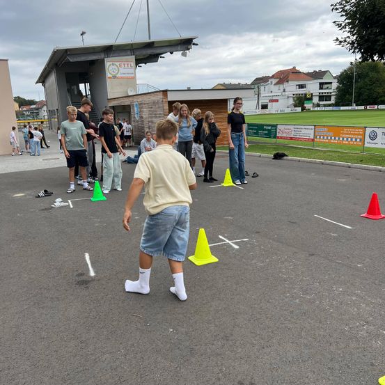 Eine Gruppe von Kindern und Jugendlichen nimmt an einem Outdoor-Agilitätstraining teil. Ein Junge führt, gefolgt von anderen. Sie navigieren durch einen Parcours, der mit Kegel und Kreide markiert ist.