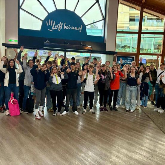 Eine Gruppe von Menschen in Freizeitkleidung posiert für ein Foto in einem Gebäude mit dem Schriftzug 'Lift bei uns!'. Einige von ihnen heben ihre Hände.