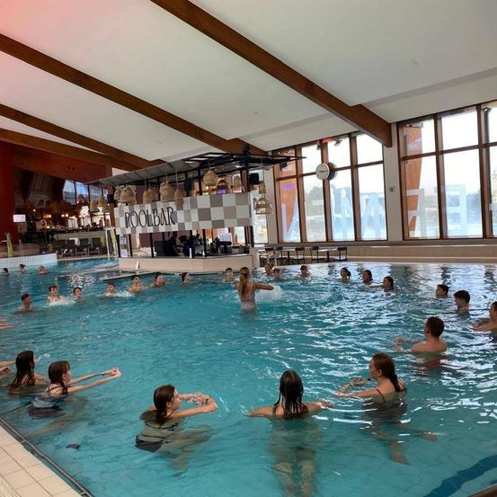 Ein Hallenbad mit einer Poolbar ist mit Menschen gefüllt, die an einem Aqua-Fitnesskurs teilnehmen. Der Pool hat klares blaues Wasser und die Decke ist aus Holz. Die Poolbar hat ein kariertes Muster.