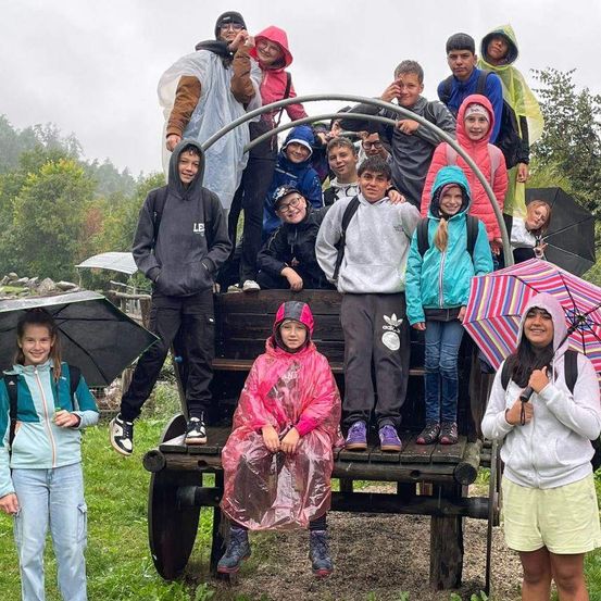 Eine Gruppe von Schülern steht in einem hölzernen Wagen mit Regenponchos und Regenschirmen und posiert für ein Foto auf einem Grasfeld mit Bäumen im Hintergrund.