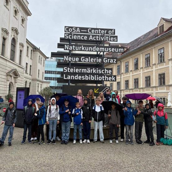 Eine Gruppe von Kindern posiert für ein Foto vor einem großen Schild vor einem Gebäude. Das Schild hat mehrere Namen und Pfeile, die in verschiedene Richtungen zeigen.