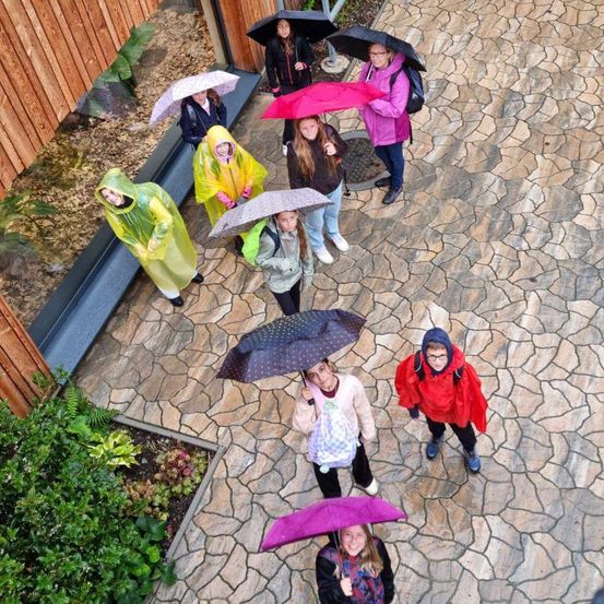 Eine Gruppe von Personen, wahrscheinlich Schüler, steht in einem Innenhof und hält alle Regenschirme, was auf Regen hindeutet. Sie tragen Regenmäntel und Stiefel.