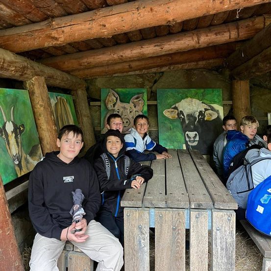 Eine Gruppe von Jungen sitzt um einen Holztisch in einer rustikalen Umgebung. Sie sind von Tiergemälden umgeben.