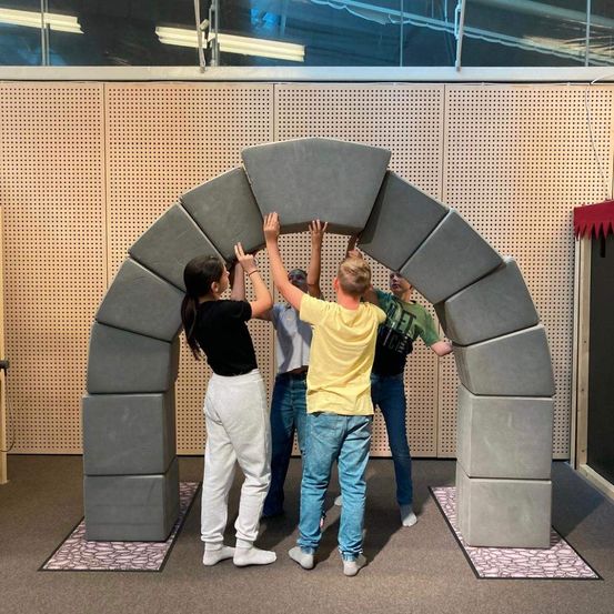 Vier Personen stehen um einen großen Bogen aus grauen Blöcken herum, jeder streckt die Hände nach oben, um die Spitze zu berühren. Der Bogen ist an einer strukturierten Wand positioniert. Die Gruppe besteht aus einer Frau in einem schwarzen T-Shirt, einem Mann in einem gelben T-Shirt, einem Mann in einem grauen T-Shirt und einer weiteren Frau in einem grünen T-Shirt.