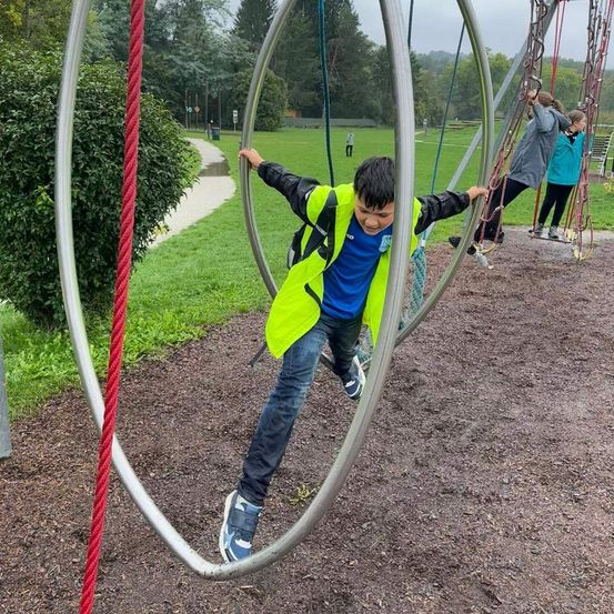 Ein junger Junge in einer gelben Weste schwingt auf einem großen Metallring in einem Park. Zwei andere Personen sind in der Nähe, eine auf einer Schaukel. Bäume und ein Weg sind im Hintergrund.