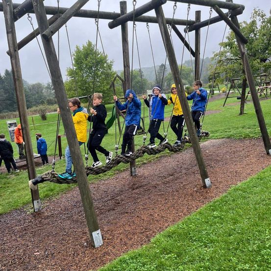 Mehrere Kinder spielen auf Schaukeln in einem Außenbereich. Sie sind in Jacken und Turnschuhen gekleidet. Einige halten sich an den Ketten der Schaukeln fest. Der Boden ist mit Schmutz und Gras bedeckt.
