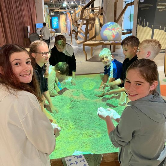 Eine Gruppe von Kindern spielt in einer Sandwanne in einem Museum, lächelt und ist in die Aktivität vertieft.