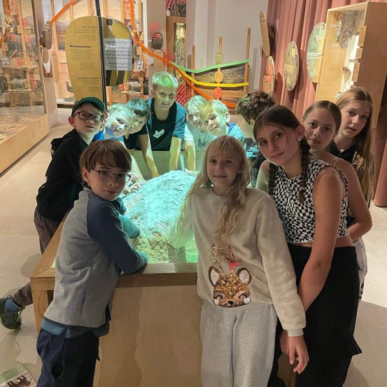 Eine Gruppe von Kindern und Jugendlichen posiert für ein Foto in einem Museum. Sie stehen um eine große blaue Ausstellung herum.