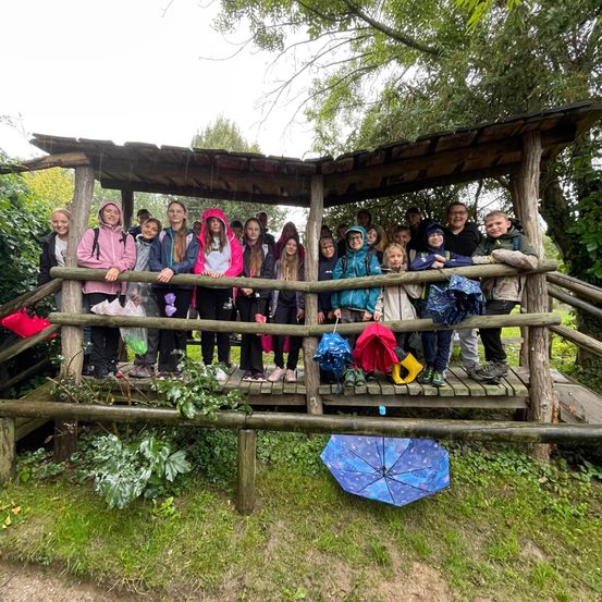 Eine Gruppe von Kindern und Erwachsenen steht in einer hölzernen Laube, hält Regenschirme und Regenmäntel und posiert lächelnd für ein Foto.