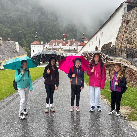 Fünf Mädchen stehen auf einer nassen Straße und halten bunte Regenschirme. Hinter ihnen steht ein Schloss und ein nebliger Berg. Sie tragen alle Jacken und Sneakers.