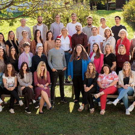 Eine große Gruppe von Menschen ist für ein Gruppenfoto in einem Park versammelt, wobei einige auf Bänken sitzen und andere dahinter stehen. Bäume und Gras sind im Hintergrund.