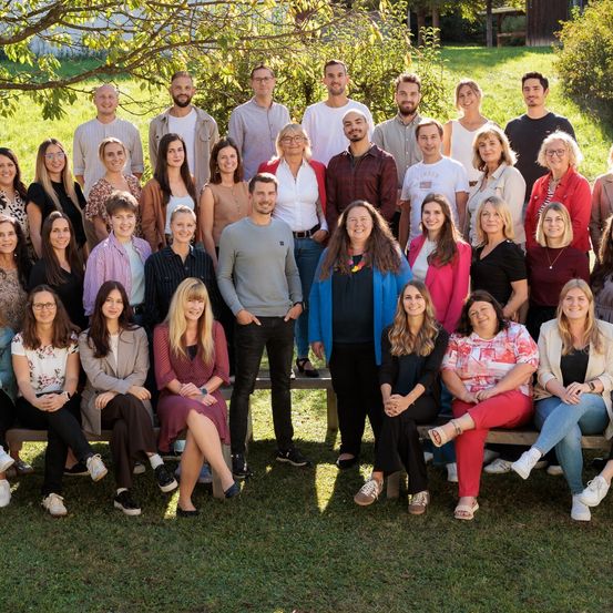 Eine große Gruppe von Menschen ist für ein Gruppenfoto in einem Park versammelt, wobei einige auf Bänken sitzen und andere dahinter stehen. Bäume und Gras sind im Hintergrund.