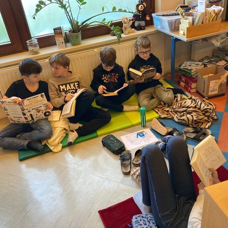 Fünf Jungen sitzen auf einer Matte am Fenster und lesen Bücher. Schuhe und ein Tisch mit Büchern und Kartons sind in der Nähe.
