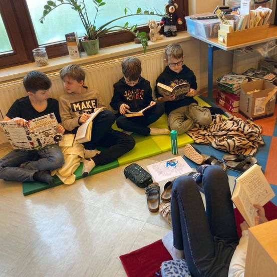 Fünf Jungen sitzen auf einer Matte am Fenster und lesen Bücher. Schuhe und ein Tisch mit Büchern und Kartons sind in der Nähe.