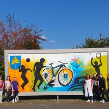 Gruppenfoto vor einem farbenfrohen Wandbild. Das Wandbild zeigt verschiedene Sportarten und Aktivitäten. Die Menschen im Bild stehen eng beieinander.