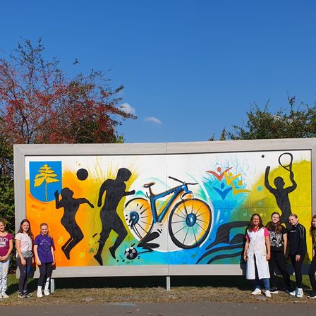 Gruppenfoto vor einem farbenfrohen Wandbild. Das Wandbild zeigt verschiedene Sportarten und Aktivitäten. Die Menschen im Bild stehen eng beieinander.