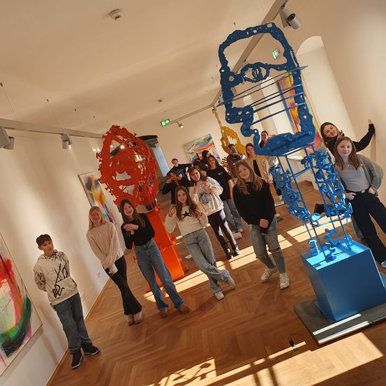 Eine Gruppe junger Leute posiert für ein Foto in einer Kunstgalerie, mit farbenfrohen abstrakten Skulpturen im Hintergrund.