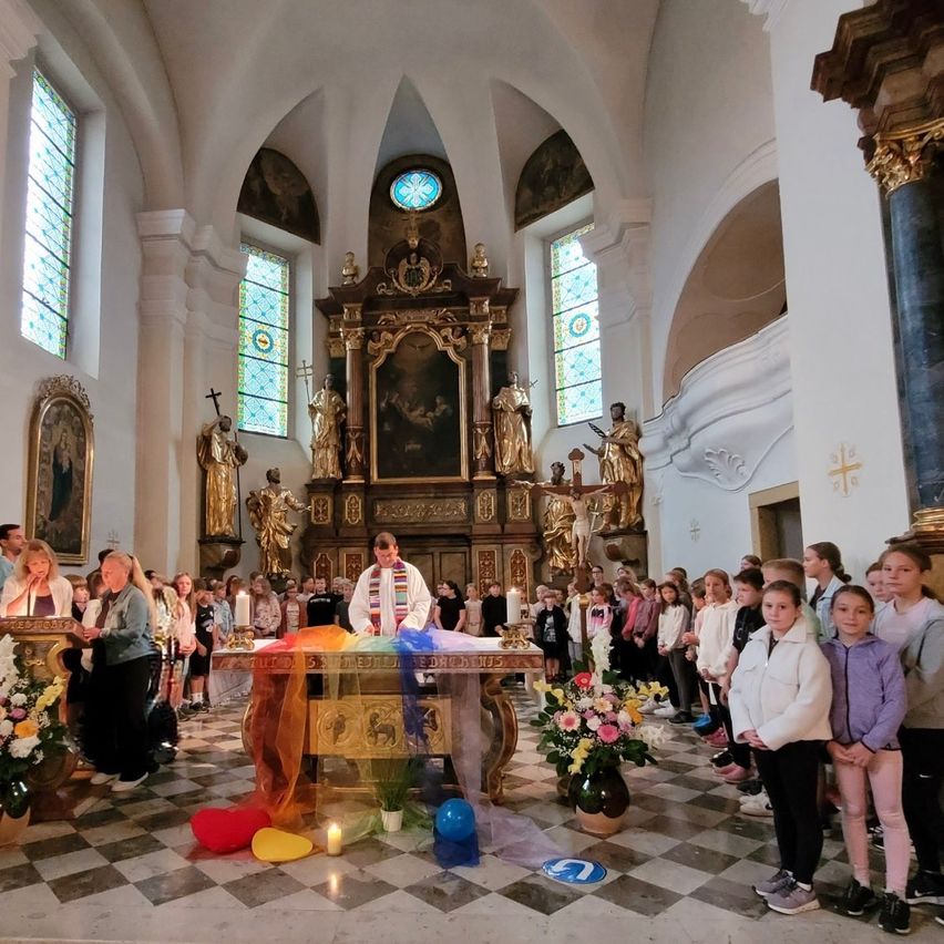 Eine religiöse Zeremonie in einer Kirche, mit einem Priester am Altar, umgeben von Kindern und Erwachsenen, die Kerzen halten. Der Altar ist mit Blumen und Luftballons geschmückt, und Statuen stehen dahinter.