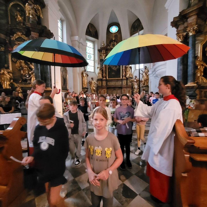 In einer Kirche versammeln sich eine Gruppe von Kindern und Erwachsenen. Zwei Erwachsene halten bunte Regenschirme. Die Kinder tragen Brillen und Turnschuhe. Der Boden ist gemustert.