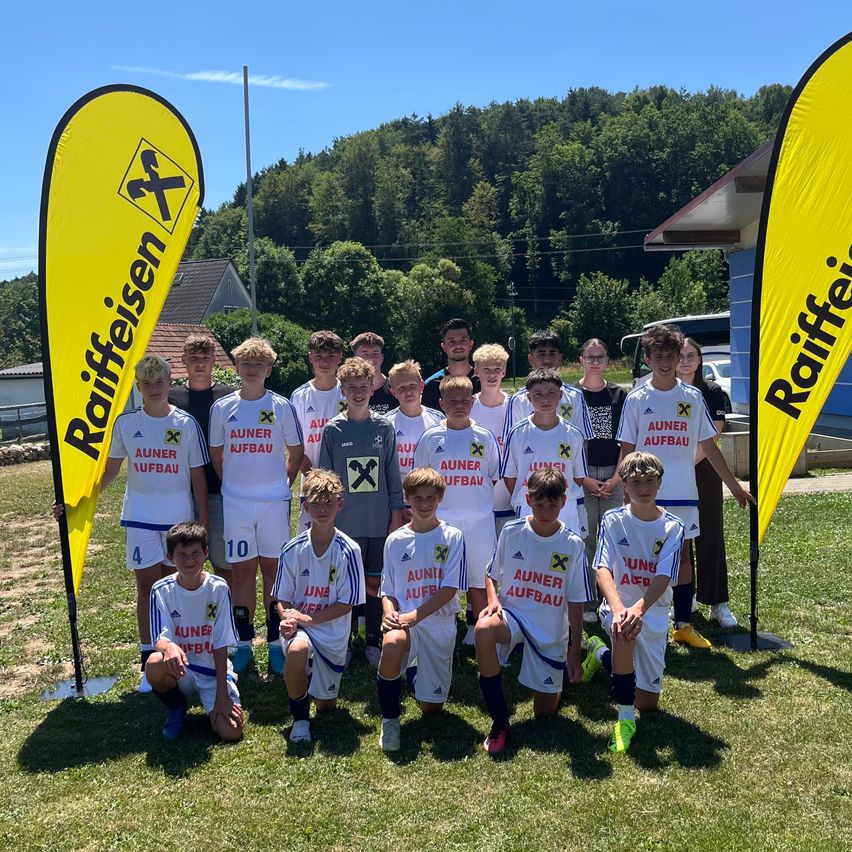 Eine Fußballmannschaft in weißen Trikots mit roten Buchstaben steht auf einem Rasenfeld. Sie posieren für ein Foto mit zwei gelben Bannern hinter ihnen. Bäume und ein Haus sind im Hintergrund.