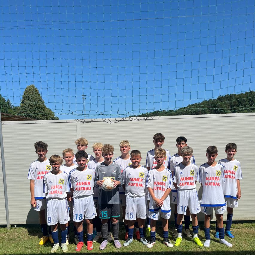 Eine Jugendfußballmannschaft posiert für ein Foto vor einem Netz. Die Spieler tragen weiße Trikots mit roten und blauen Besätzen. Die Trikots haben verschiedene Nummern auf der Rückseite.