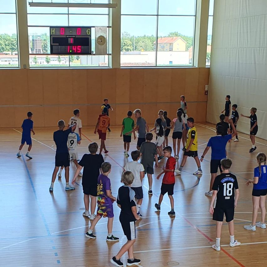 Eine Gruppe von Kindern in Trikots mit Nummern spielt ein Spiel auf einem Innenfeld. Die Anzeigetafel zeigt Unentschieden mit 0-0. Die Kinder tragen Turnschuhe.