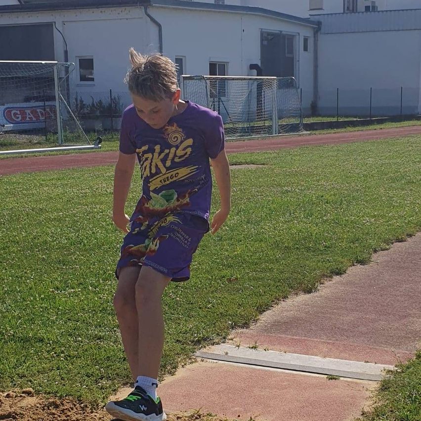 Ein junger Junge in einem lila T-Shirt steht auf einer Sandgrube auf einer Laufbahn und schaut nach unten. Das Gebiet ist von grünem Gras und einer roten Bahn umgeben. Im Hintergrund befindet sich ein Gebäude mit einem Schild, auf dem 'GAD' steht.