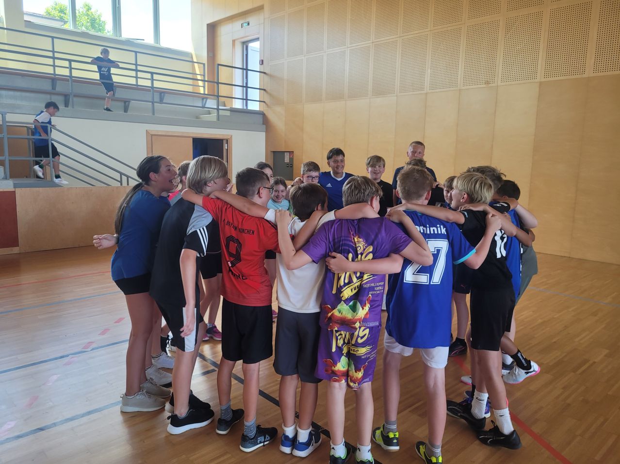 Eine Gruppe junger Menschen steht in einem Kreis im Inneren einer Turnhalle. Einige tragen Sporttrikots, andere Freizeitkleidung. Sie alle stehen sich gegenüber und machen möglicherweise eine Teambuilding-Übung. Eine Person befindet sich auf dem Balkon oben.