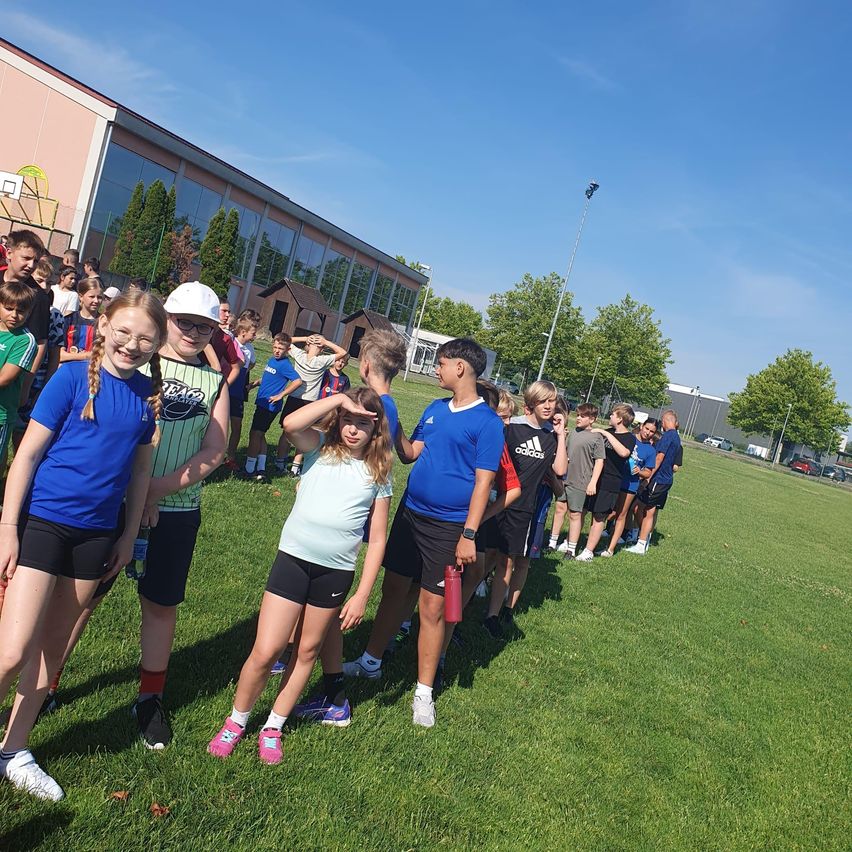Eine Gruppe von Kindern steht in einer Reihe auf einem Rasenfeld. Sie tragen Sportkleidung. Einige von ihnen lächeln, während andere etwas betrachten. Hinter ihnen befindet sich ein Gebäude mit Glasfenstern und einem Basketballplatz.